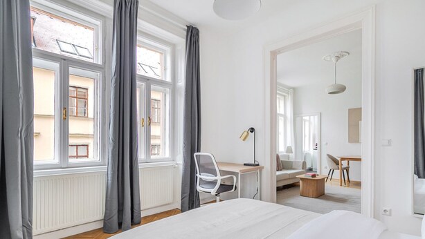 Ein modernes Hotelzimmer mit einem großen Bett, einem Schreibtisch mit Stuhl, einem kleinen Tisch und Blick auf die Straße durch große Fenster.