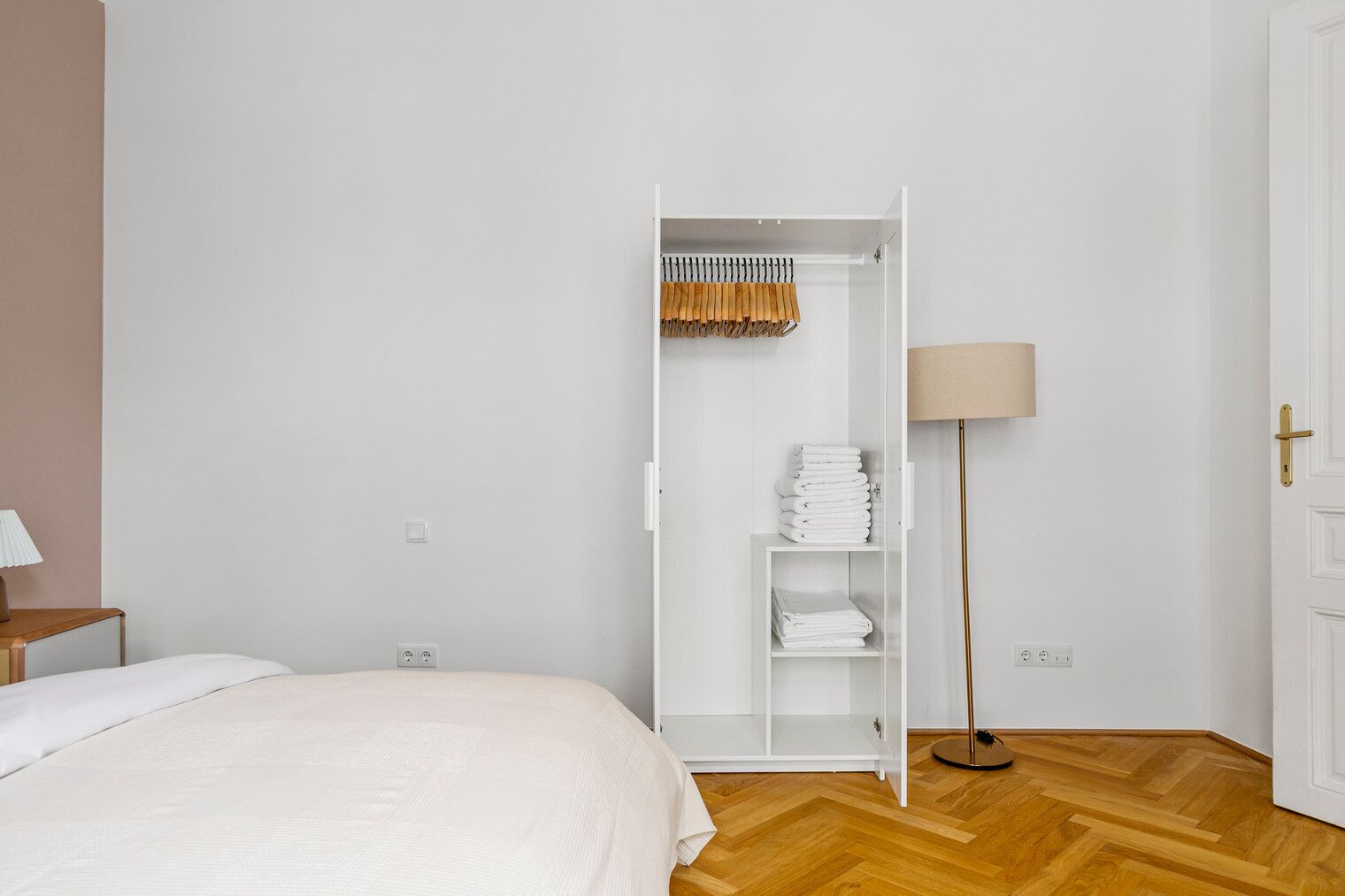 Une chambre à coucher avec un lit, une armoire blanche contenant des vêtements pliés, une lampe sur pied et une porte.