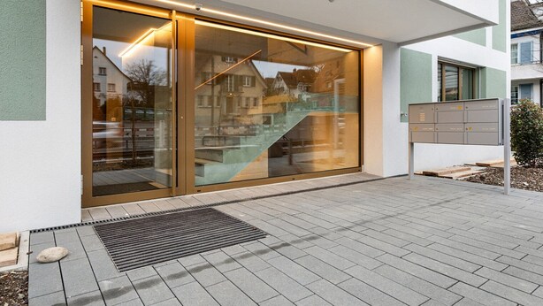 A modern building entrance with a glass door, a metal mailbox, and a paved area with a drainage grate. - Blueground | Zollikon, terrace, nr Zurich Zoo (Zurich)