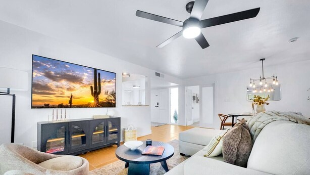 A modern living room with a ceiling fan, a large TV, and a dining area with a chandelier. - Revolta Fairway Retreat (Mesa)