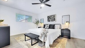 A modern bedroom with a bed, bedside tables, a bench, a ceiling fan, and a large window. - Revolta Fairway Retreat (Mesa)