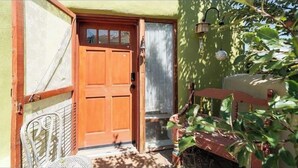 A wooden door with a glass panel, a metal chair, and a small table outside a building with a green wall and a hanging light fixture.