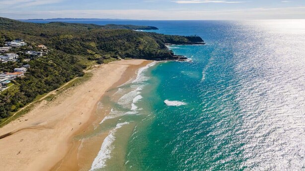 Une vue aérienne d ’une plage, avec une mer d’un bleu limpide, un rivage sablonneux et une zone boisée où se trouvent des bâtiments en arrière-plan.