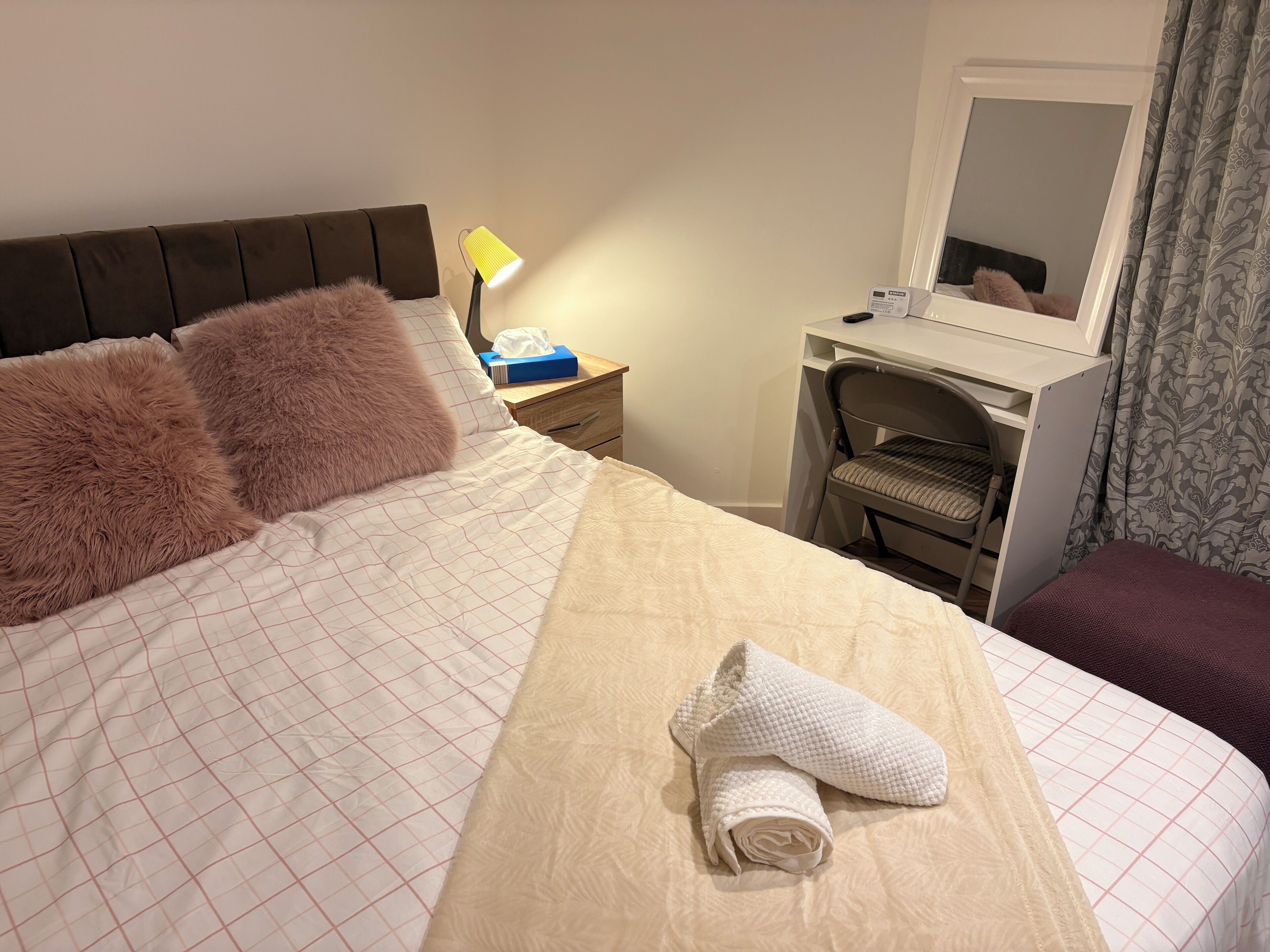 A hotel room with a bed, a desk, a chair, a mirror, and a bedside table with a lamp.