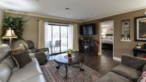 Ein Wohnzimmer mit Sofa, Sessel, Couchtisch und Fernseher. Ein Fenster mit Vorhängen, eine Pflanze und eine Tür zu einem anderen Raum sind ebenfalls vorhanden.