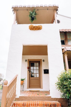 Un bâtiment blanc avec une porte marron, un petit porche avec des marches et une plante suspendue au-dessus de la porte.