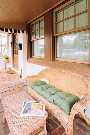 A wicker sofa with a green cushion and a magazine on a matching ottoman, a brick floor, and a window with blinds.