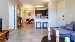 A modern living room with a blue sofa, two barstools, a dining table with red chairs, a kitchen area, and a hallway leading to a door. - Modern 1BR Condo w/ Pool & BBQ Near Fort Myers (Fort Myers)