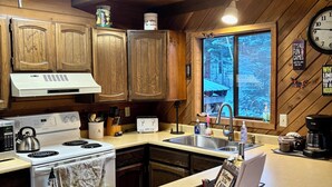 A kitchen with wooden cabinets, a stove, a sink, and a window with a view of trees. - Cozy Cabin in the Woods, Anatone, WA (Anatone)