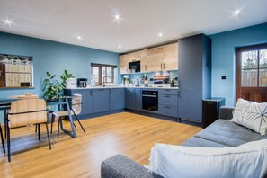 A modern kitchen with blue cabinets, a dining area with a wooden table and chairs, and a grey sofa. - The Old Milking Parlour - 1 Bed Cottage - Amroth (Amroth)