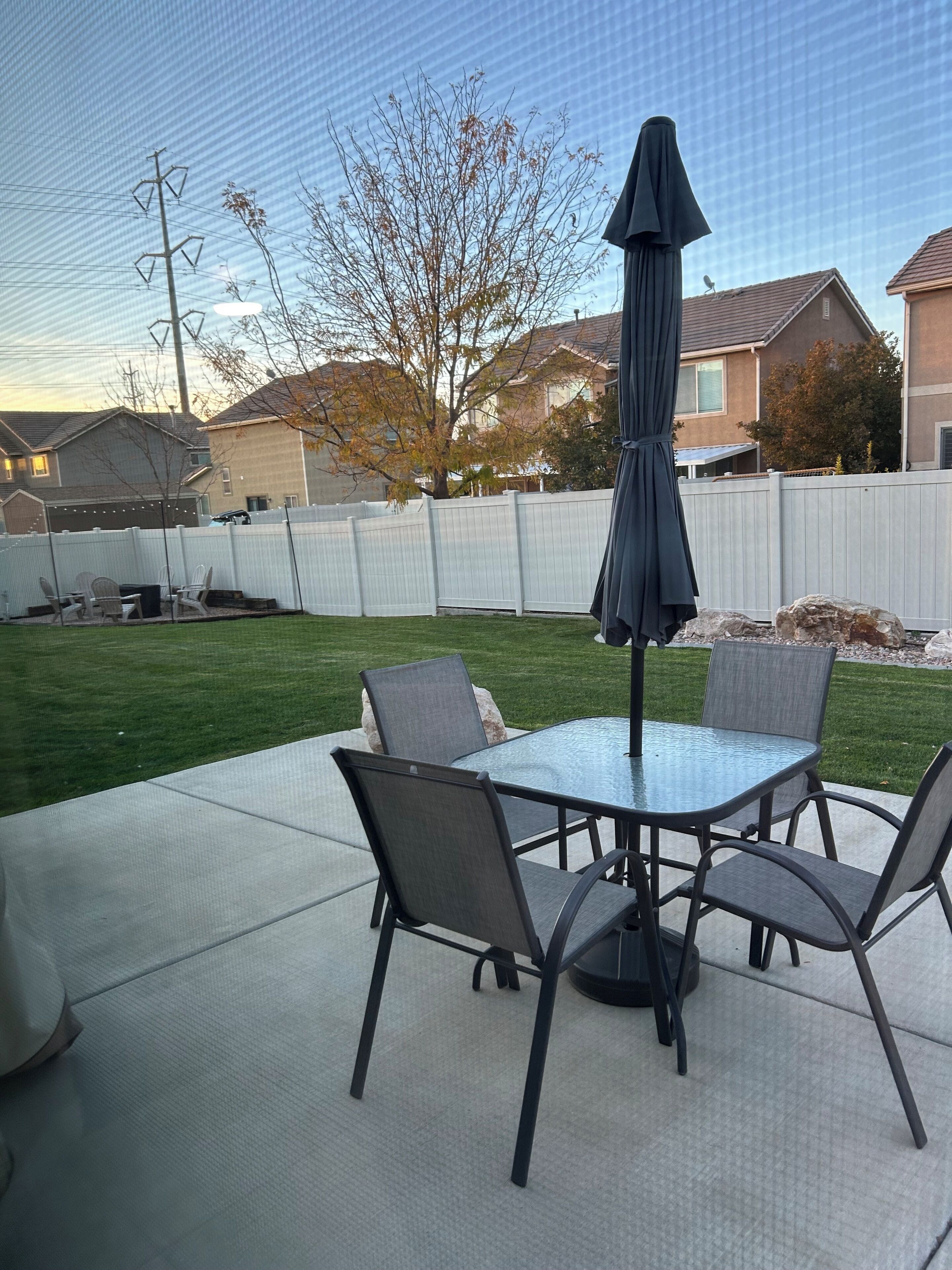 An outdoor patio with a glass table and four chairs, a dark umbrella, and a white fence in the background.