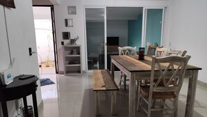 Zona pranzo con tavolo in legno e panche, un tavolino laterale con una lampada, e una porta scorrevole in vetro che dà su un'altra stanza.