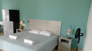 Un letto rifatto con una coperta azzurra, due asciugamani arrotolati, un comodino con una lampada e una bottiglia d'acqua, un piccolo mobiletto con una ventola e un'unità di storage a parete.