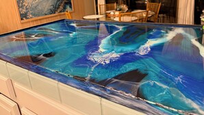 Een zwembad met glazen rand, een blauw-wit marmeren ontwerp, houten meubels en een eethoek op de achtergrond.