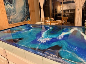 A glass-top pool with a blue and white marble design, wooden furniture, and a dining area in the background. - Oceanfront Artist's Condo Panoramic Maui Sunsets (Kihei)