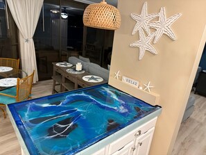 A room with a glass-top table, wicker chairs, a hanging light fixture, and decorative starfish on the wall. - Oceanfront Artist's Condo Panoramic Maui Sunsets (Kihei)