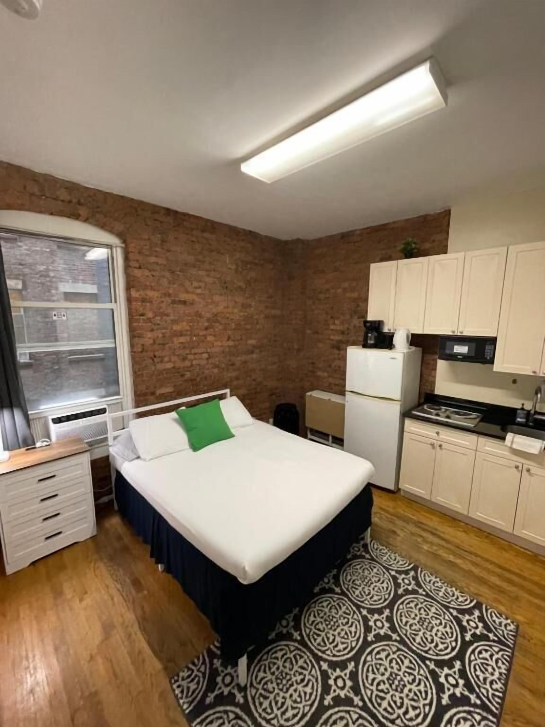 A compact bedroom with a bed, a small desk, a window, a kitchenette, and a patterned rug.