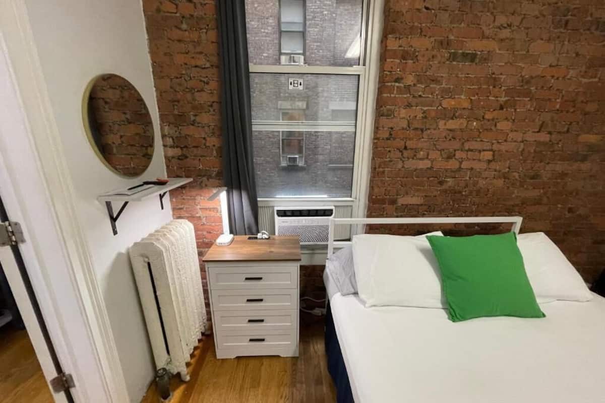 A bedroom with a brick wall, a window with blinds, a radiator, a bedside table with a lamp, a bed with white and green pillows, and a circular mirror on the wall.
