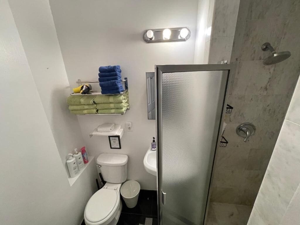 Une petite salle de bain comprenant des toilettes, un lavabo et une douche. Des serviettes sont pliées et empilées sur une étagère, et une poubelle est visible.