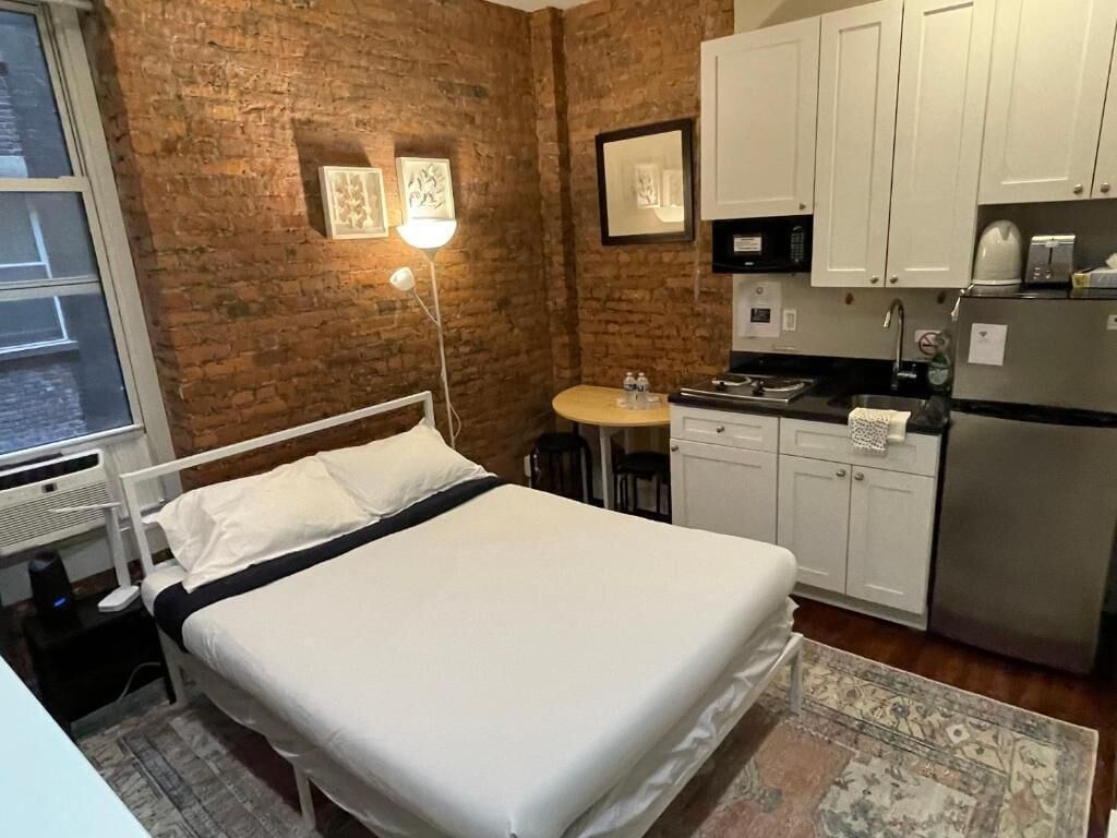 Un dormitorio con cama, mesita de noche, lámpara, una pequeña mesa redonda con sillas y una kitchenette con armarios y electrodomésticos.