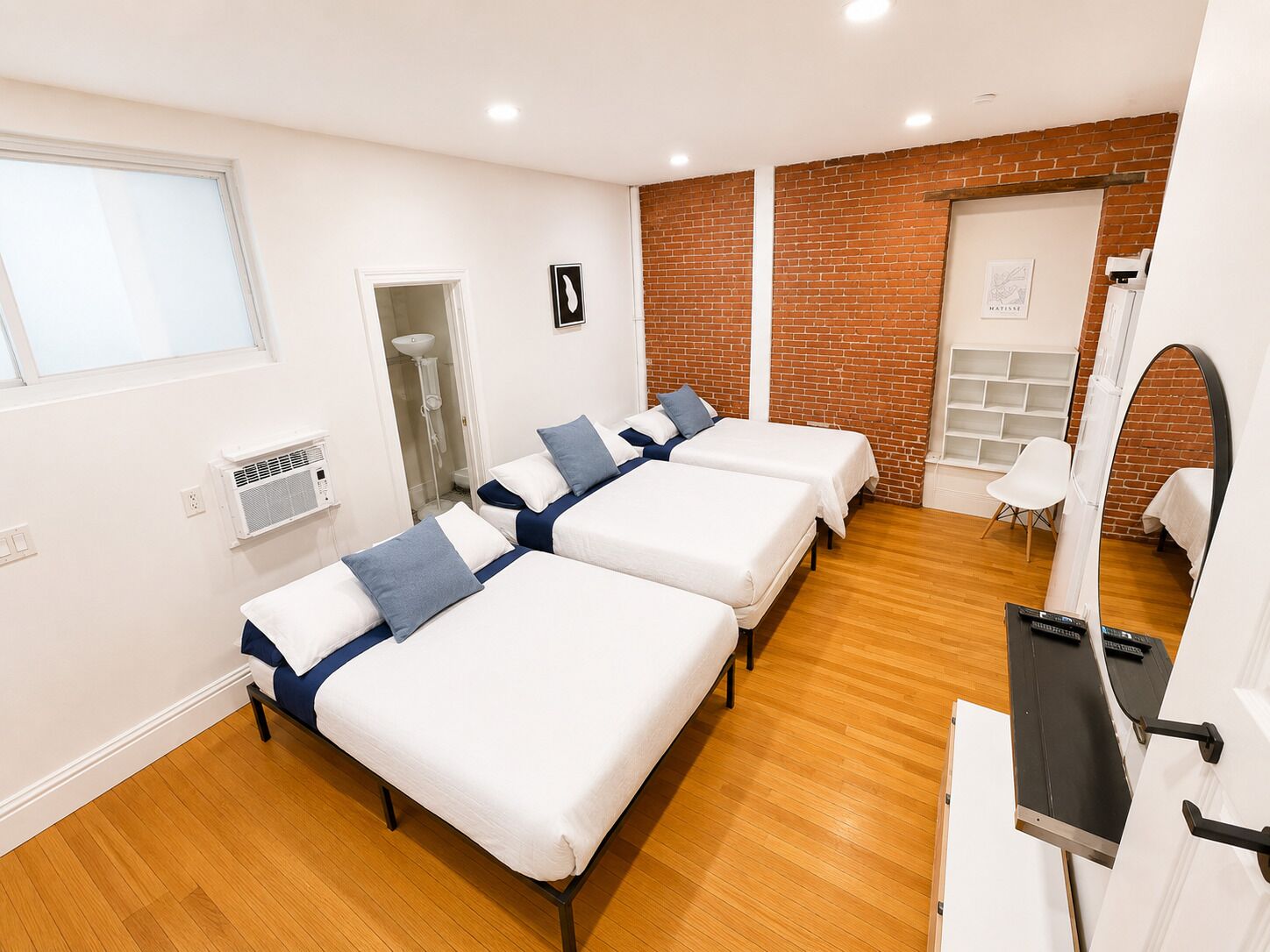 Ein Hotelzimmer mit drei Betten, einem kleinen weißen Stuhl, einem Spiegel und einer Ziegelwand.