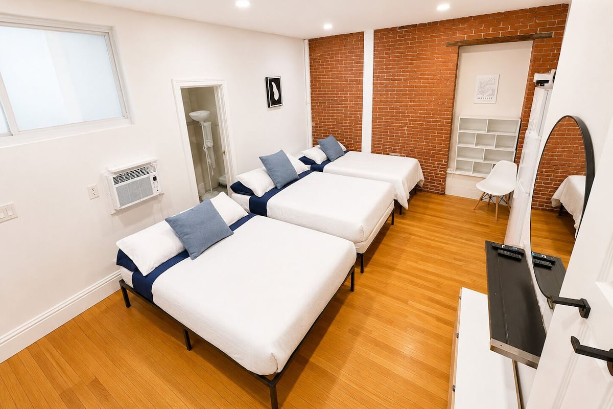 A hotel room with three beds, a small white chair, a mirror, and a brick wall.