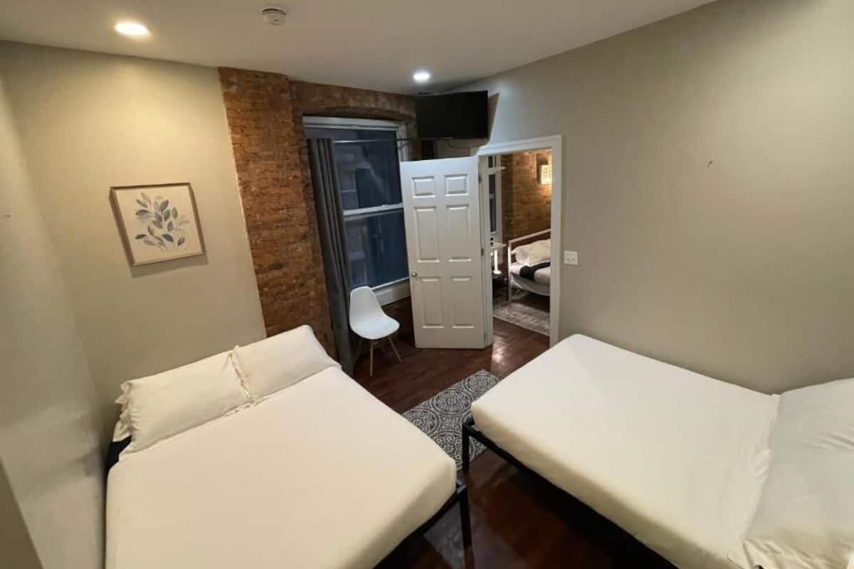 A small, well-lit room with two single beds, a white chair, a door leading to another room, and a TV mounted on the wall.