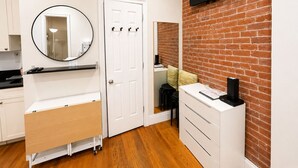 A compact hotel room with a kitchen area, a white wardrobe, a flat-screen TV mounted on a brick wall, and a round mirror. - Nest Aparthotel Manhattan  (New York)