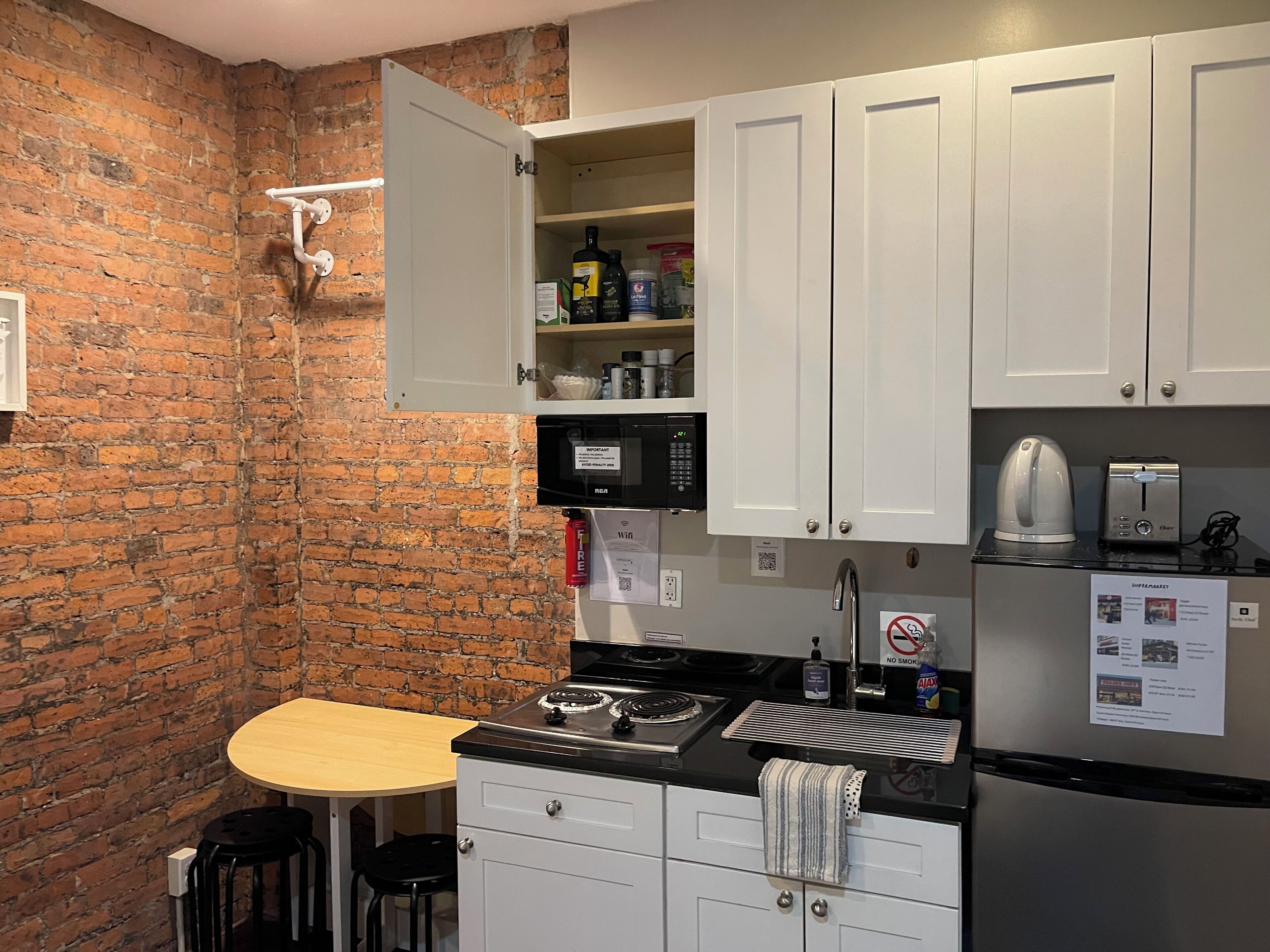A kitchen with a brick wall, white cabinets, a microwave, a kettle, and a refrigerator.
