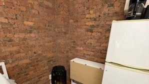 A small, narrow room with a brick wall, a white bed, a small stool, a white refrigerator, and a coffee maker on the wall. - Nest Aparthotel Manhattan  (New York)