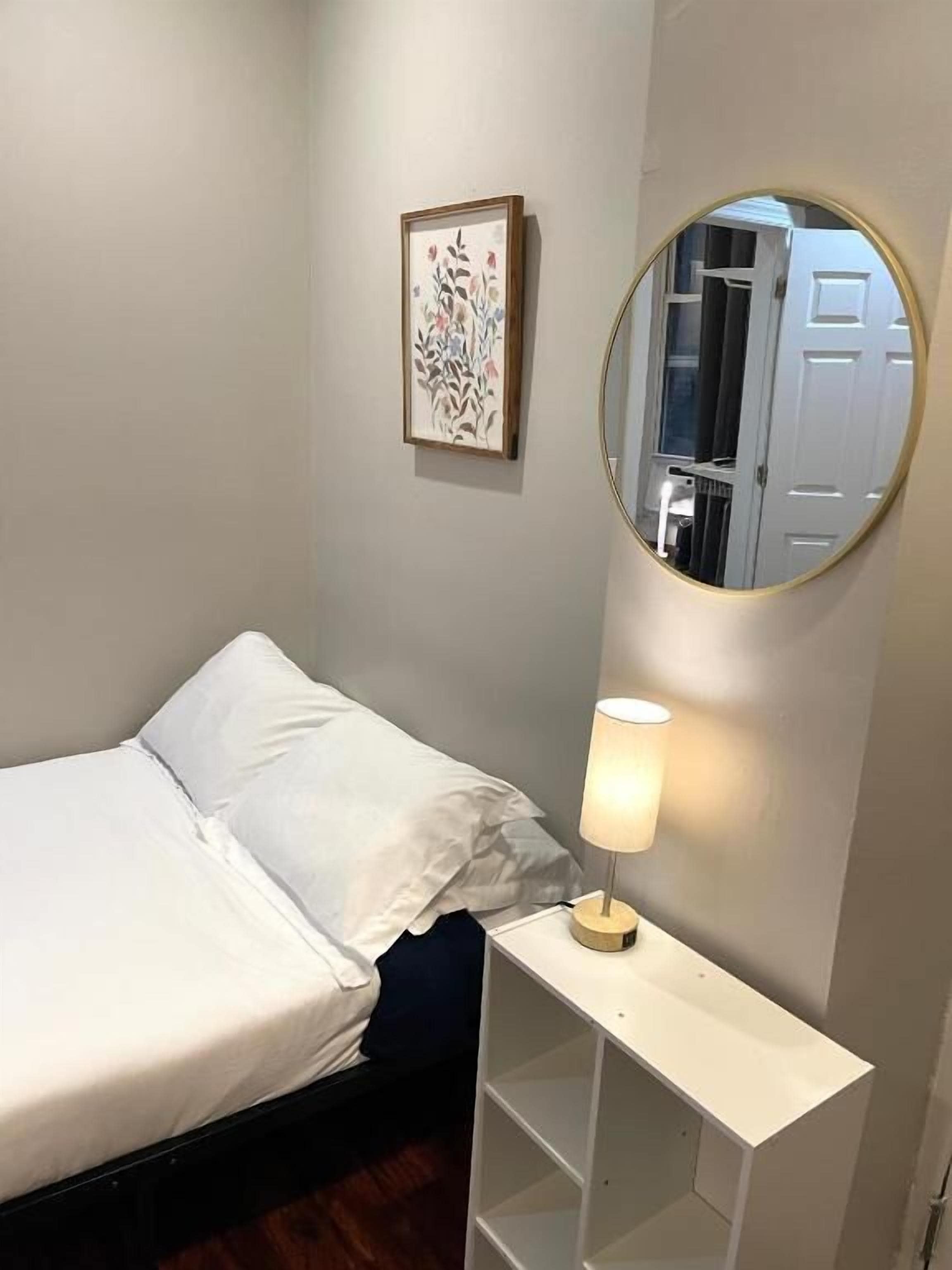 A neatly made bed with white linens, a bedside lamp, a framed floral artwork on the wall, and a round mirror on a white console table.