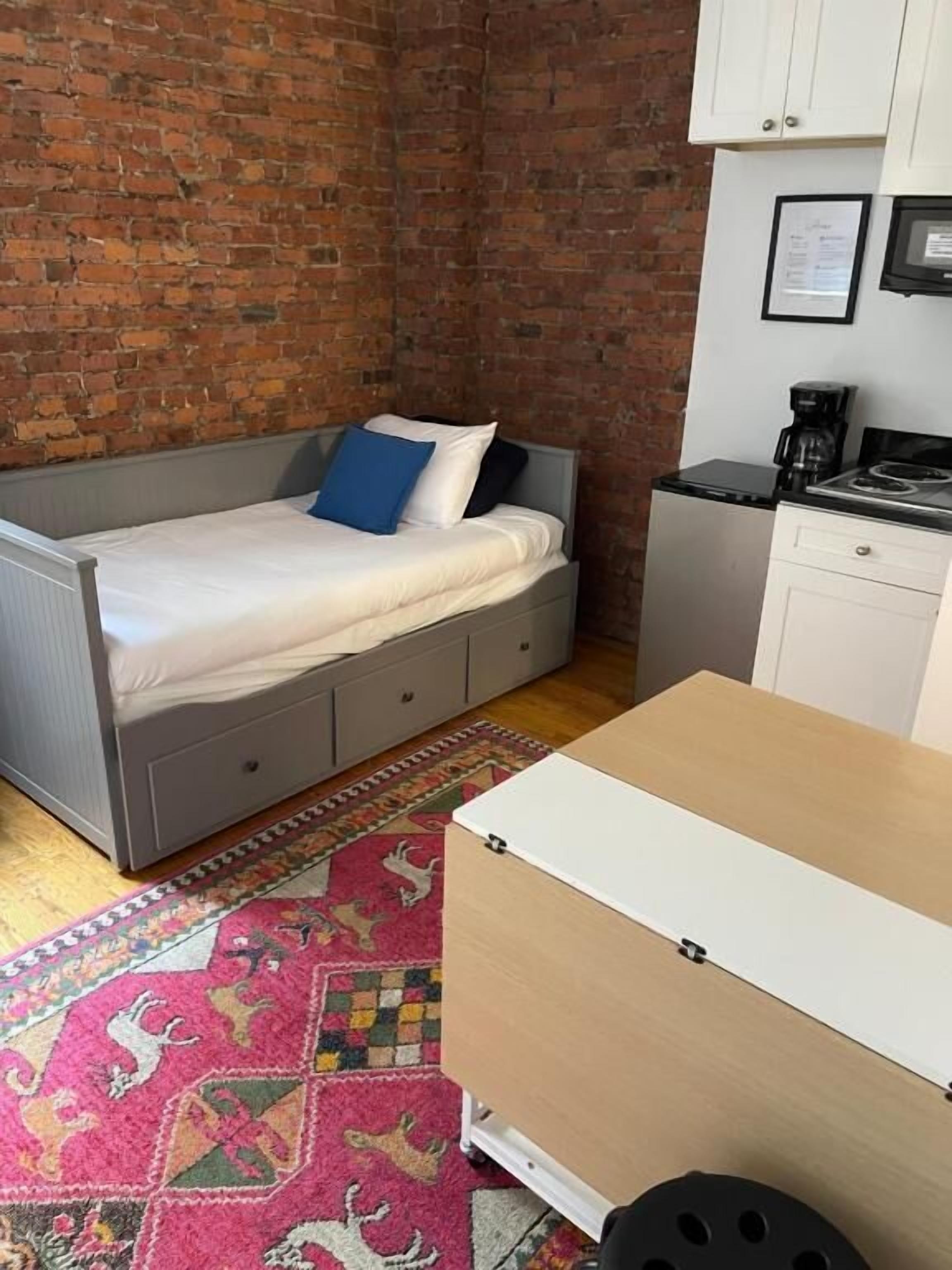 A compact room with a bed, a small kitchenette, and a patterned rug.