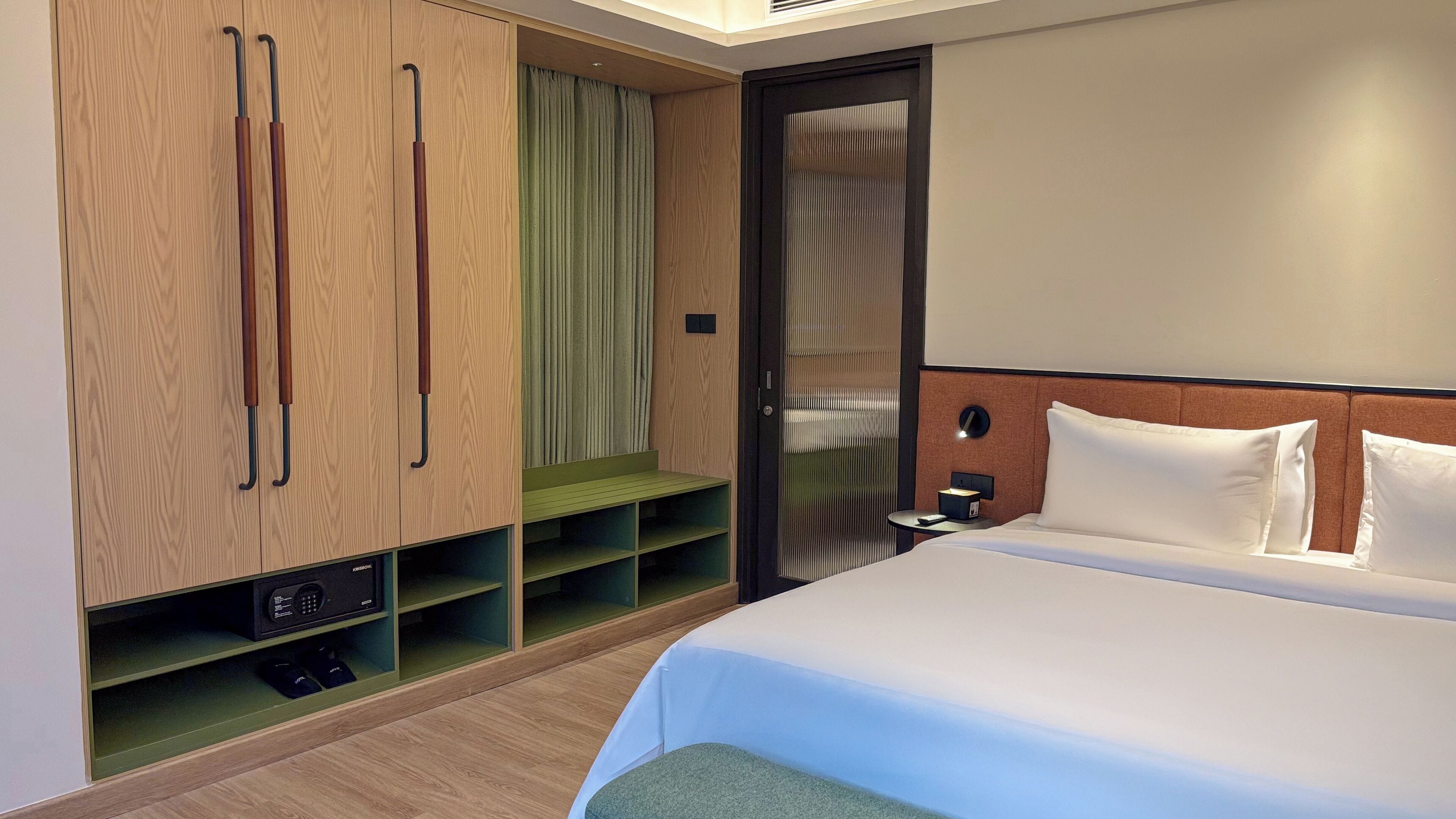 A modern hotel room with a bed, a nightstand, a wardrobe with a mirror, and a seating area with a bench.