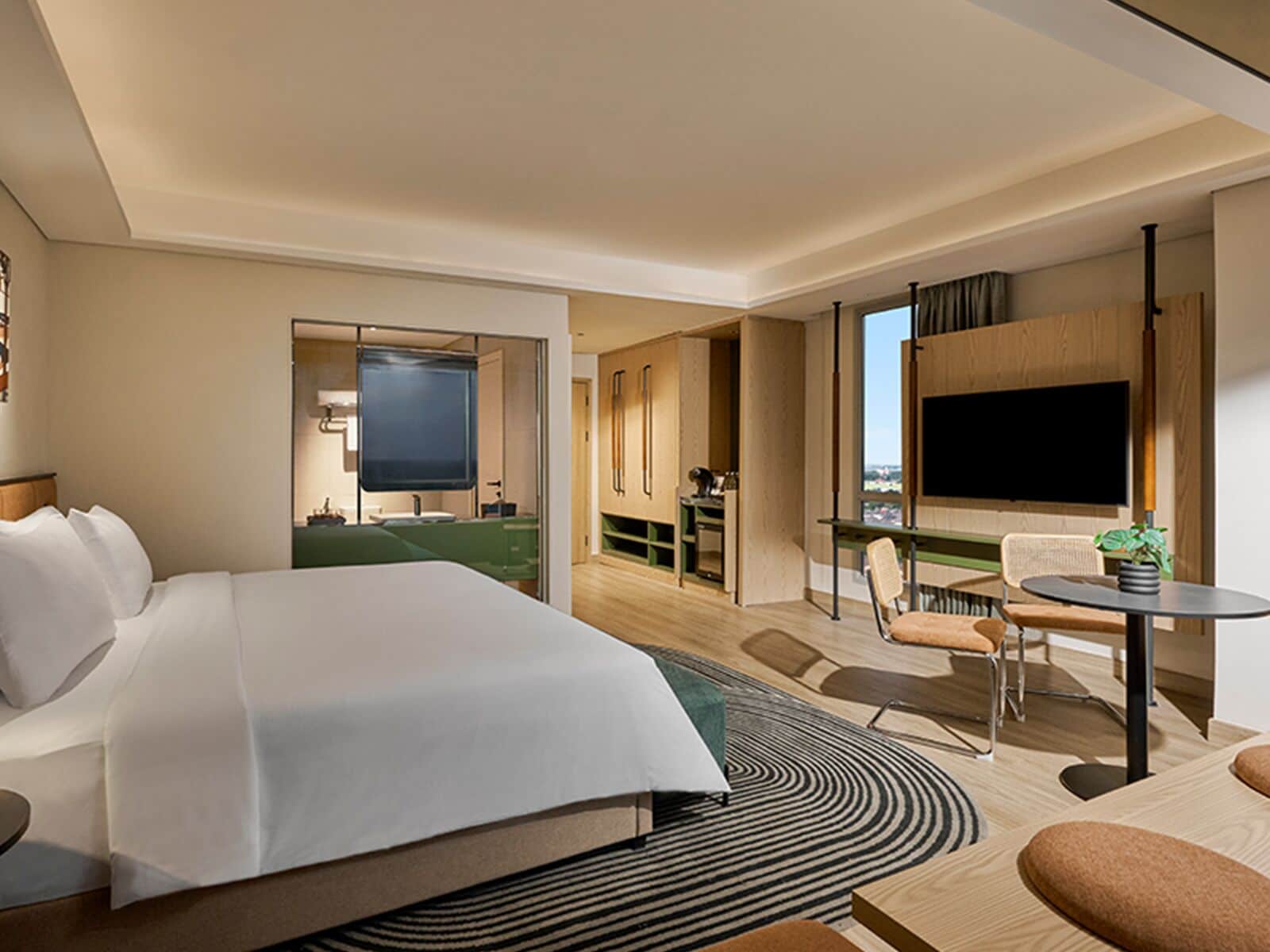 A hotel room with a large bed, a desk, a chair, a TV, and a view of the city.