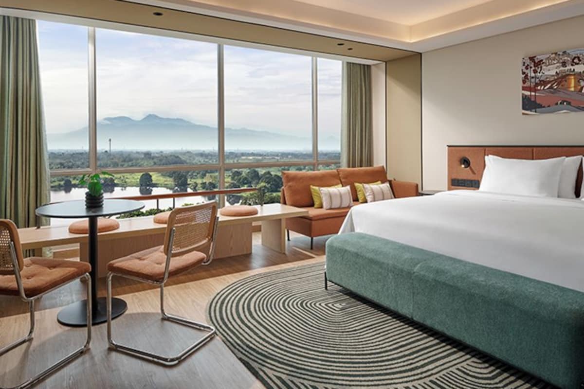 Habitación de hotel con una cama grande, una mesa de comedor con sillas, un sofá y vistas a un paisaje montañoso a través de amplias ventanas.