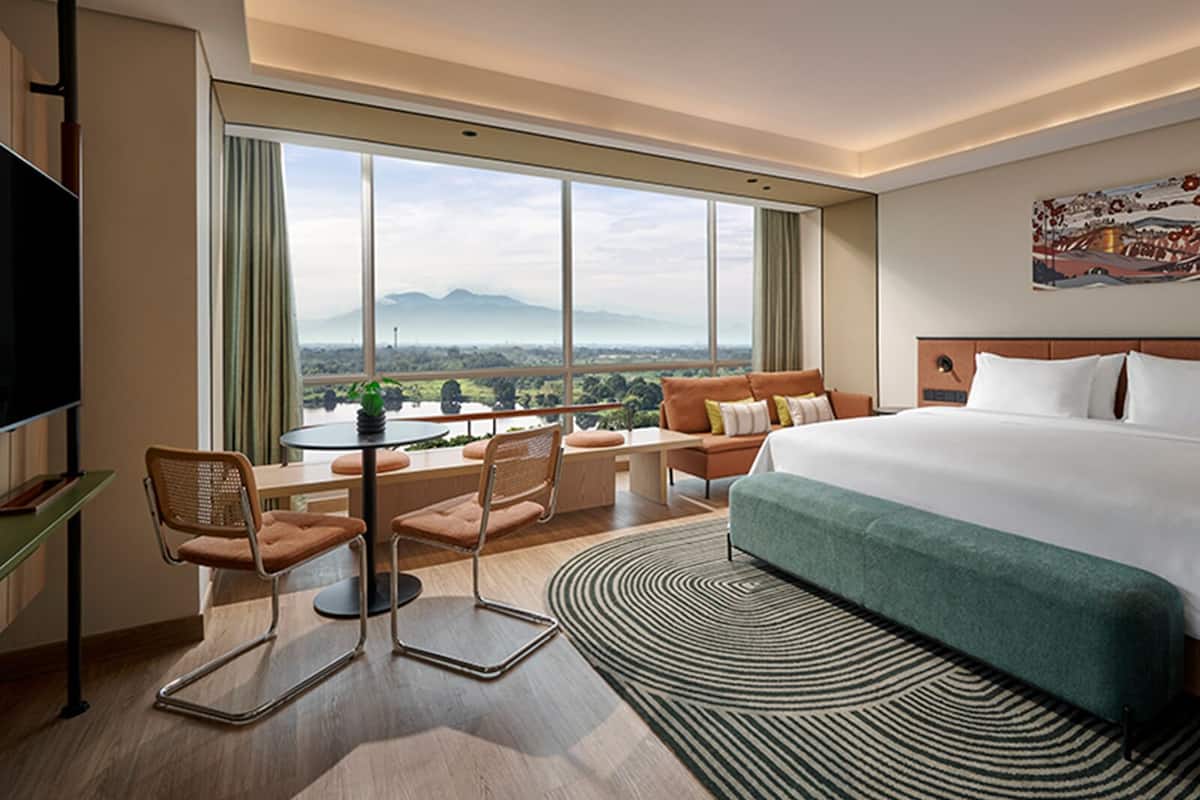 A hotel room with a large bed, a desk with a chair, a view of mountains, and a painting on the wall.