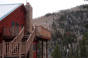 Una baita di legno con camino in pietra, un balcone in legno e vista su una montagna boscosa.