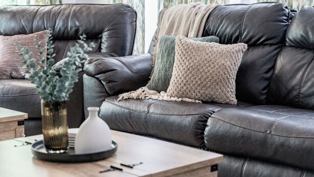 Eine dunkelbraune Ledersofa mit gewebter Tagesdecke und einem strukturierten Kissen. Auf einem Holznachttisch neben dem Sofa steht eine kleine weiße Vase mit Grünpflanzen.