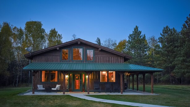 A two-story log cabin with a green metal roof, a covered porch with chairs, and a visible front door. - Secluded Cabin near Lake Vermillion, Sleeps 10, with Hot Tub, and Pet Friendly (Angora)