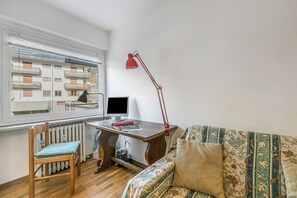 Ein Zimmer mit Holzboden, ein Holzstuhl mit Kissen, eine Couch mit gemustertem Bezug, eine rote Schreibtischlampe, ein Computermonitor und ein Fenster mit Blick auf Gebäude.