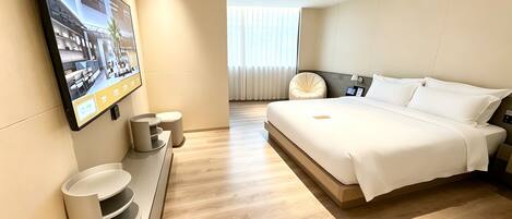 A modern hotel room with a large bed, a flat-screen TV mounted on the wall, a small seating area with a chair and a table, and a view of the hotel lobby through a window.