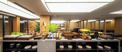 Zona de desayuno de hotel moderna con un mostrador central que exhibe alimentos, una máquina de café y una variedad de platos.