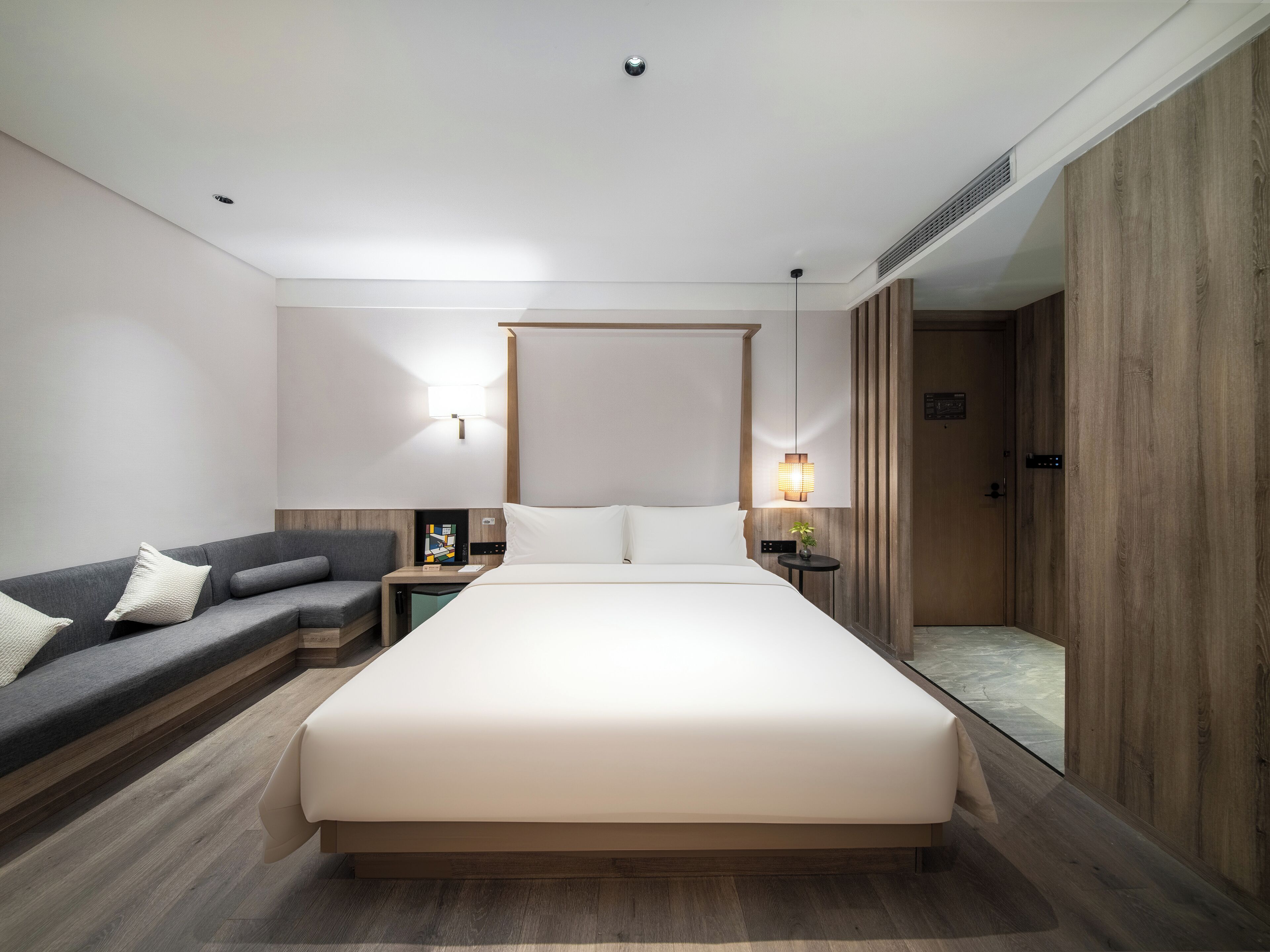 A modern hotel room with a large bed, a sofa, a bedside table, and a small table with a lamp.