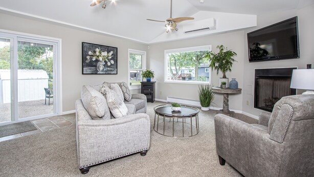 A living room with a grey sofa, a round coffee table, and a fireplace. There are ceiling fans, a TV, and a large window. - Fury House • Fenton/St Louis/Lindbergh • 4BR/3BA (Fenton)