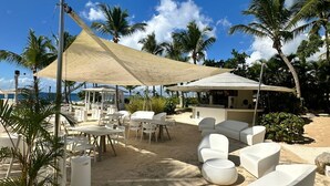 Un área de estar al aire libre frente a la playa, con mobiliario blanco, palmeras y un cielo azul despejado.