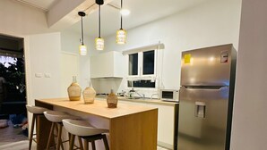 Una cocina moderna con una isla central, taburetes de bar, luces colgantes, un refrigerador de acero inoxidable y una ventana con vistas al exterior.