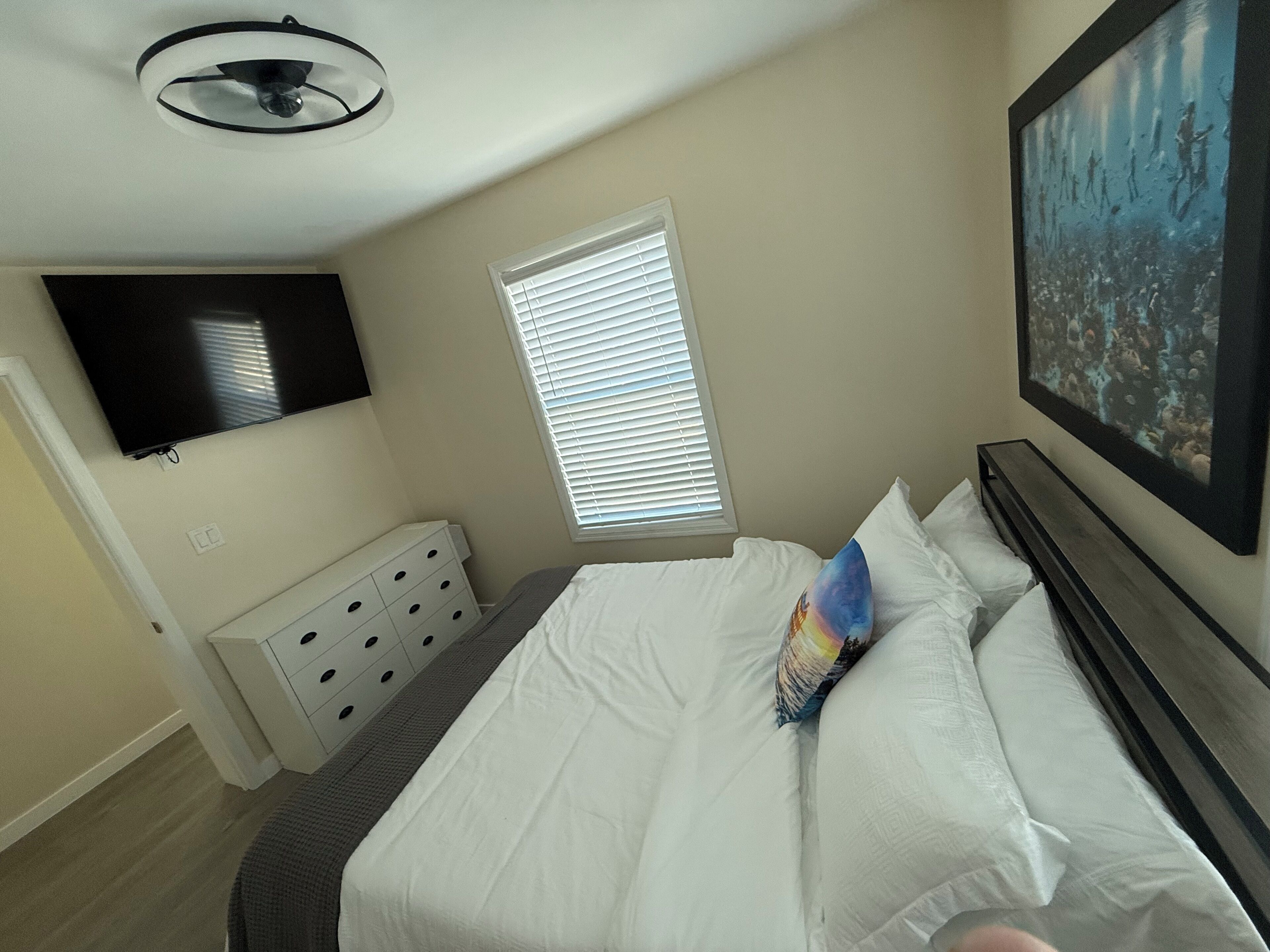 Ein Schlafzimmer mit einem Bett, einem an der Wand montierten Fernseher, einem Fenster mit Jalousien und einem Deckenventilator.