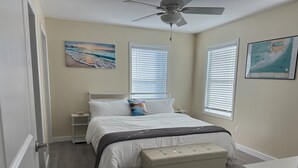 Ein Schlafzimmer mit Bett, Deckenventilator, Nachttisch, Bank und zwei Fenstern mit Jalousien.