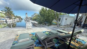 Eine Strandlounge mit Liegestühlen und einer Zierdecke, mit Blick auf einen Yachthafen und eine Statue.