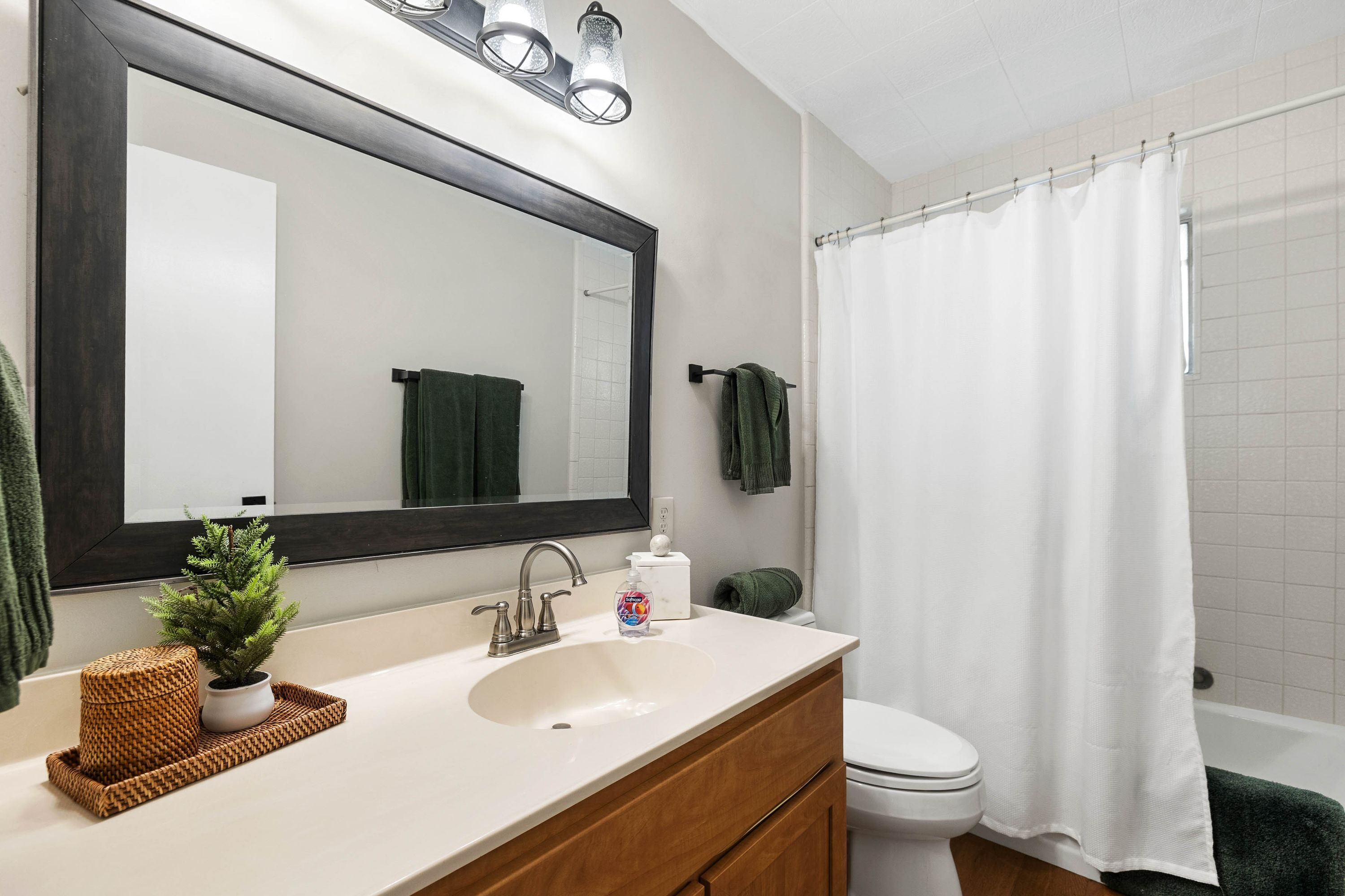 A bathroom with a large mirror, white sink, wooden vanity, green towels, and a white shower curtain.